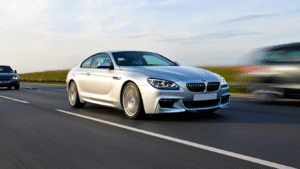 A silver 2012 BMW 640d coupe drives at speed on a UK motorway. The car gleams in the sunlight with polished reflections, its wheels and background slightly motion-blurred to convey movement. Other modern cars appear in the background, but the BMW remains the visual focus. The scene includes green roadside embankments and a partly cloudy sky.