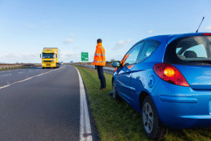 Blue hatchback broken down on M20 hard shoulder in Kent, with hazard lights on as a recovery truck approaches and driver waits safely behind barrier.