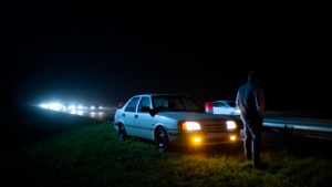 A realistic nighttime roadside scene in Kent showing a car stopped safely with hazard lights and headlights on. Traffic passes in the distance. The driver is standing well away from the vehicle behind a barrier. Dark environment with realistic lighting, documentary style, unstaged.